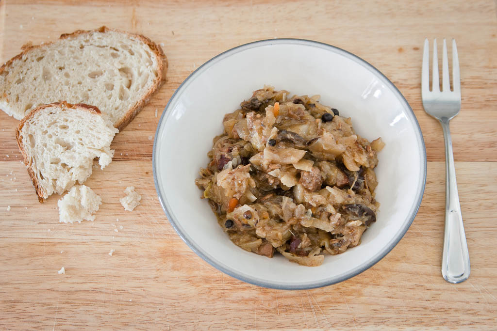 A bowl of traditional Polish bigos, a savory dish made from sauerkraut, meat, and mushrooms, served with slices of bread on a wooden table.