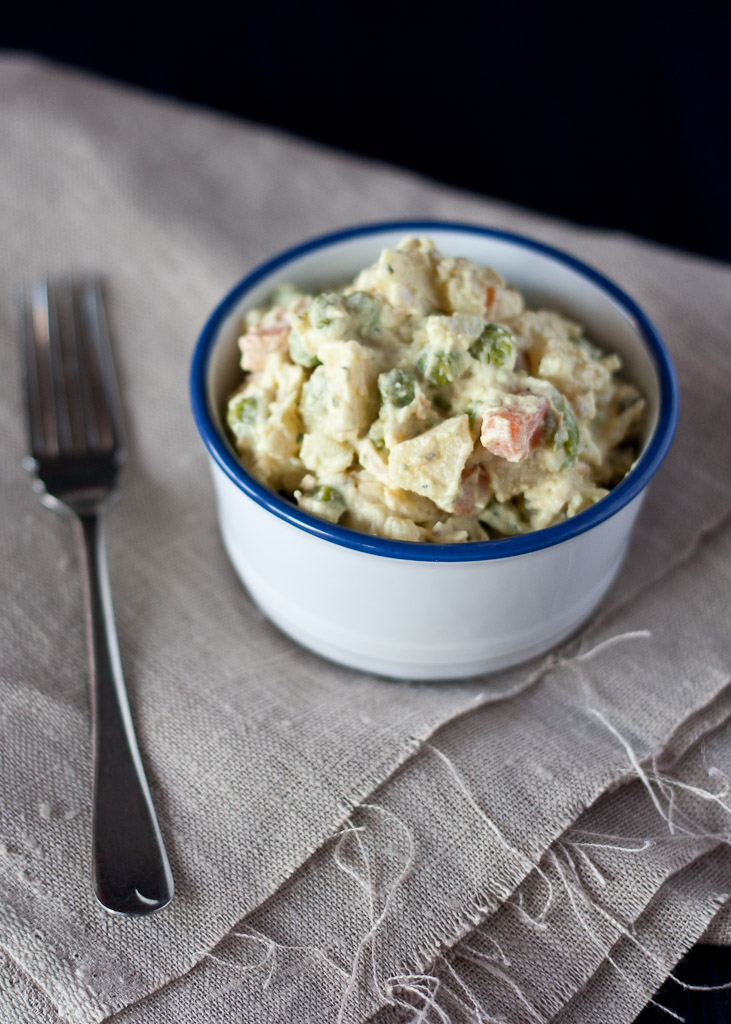 A bowl of Russian salad, featuring diced vegetables and creamy dressing, placed on a textured linen surface with a fork beside it.