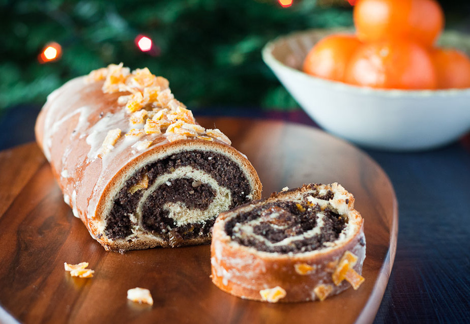 A sliced Polish poppy seed roll (makowiec) on a wooden board, showcasing its rich, nutty filling and a glaze on top, with mandarin oranges in the background.