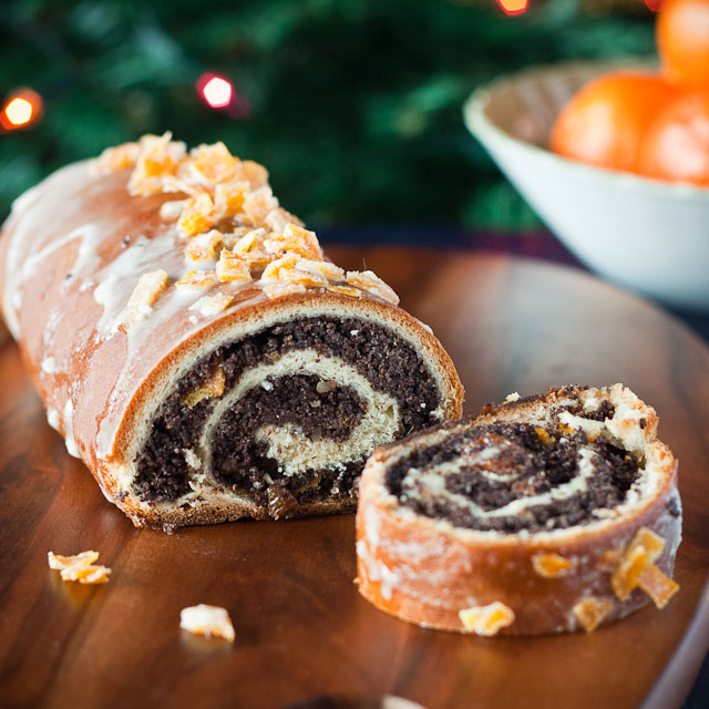 A slice of poppy seed roll (Makowiec) with a swirl of sweet poppy seed filling and a light glaze, garnished with orange zest, displayed on a wooden platter.