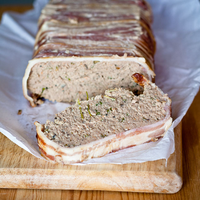 A loaf of Polish pate, wrapped in bacon, with a slice cut revealing a creamy, herb-filled interior, placed on a wooden board.