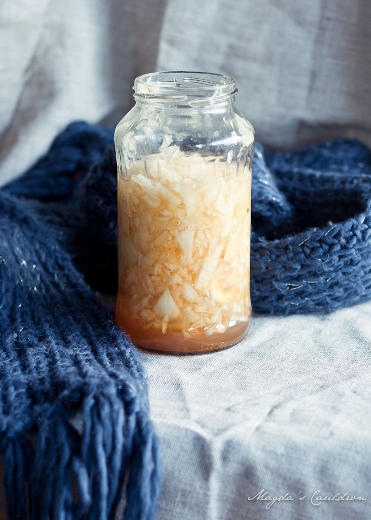 A glass jar filled with homemade onion syrup, showing chopped onions submerged in a sweet liquid, placed on a textured fabric backdrop.