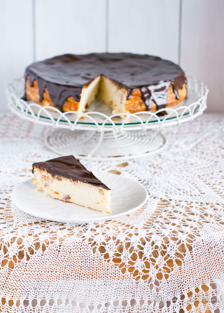 A slice of decadent Polish cheesecake topped with chocolate, served on a plate next to a whole cheesecake on a decorative stand, with a lace tablecloth in the background.