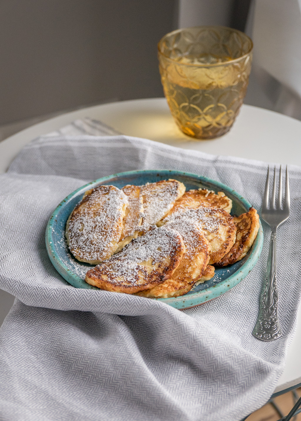 Plate of cheese pancakes dusted with powdered sugar, served with a glass of beverage in the background and a fork beside the plate.