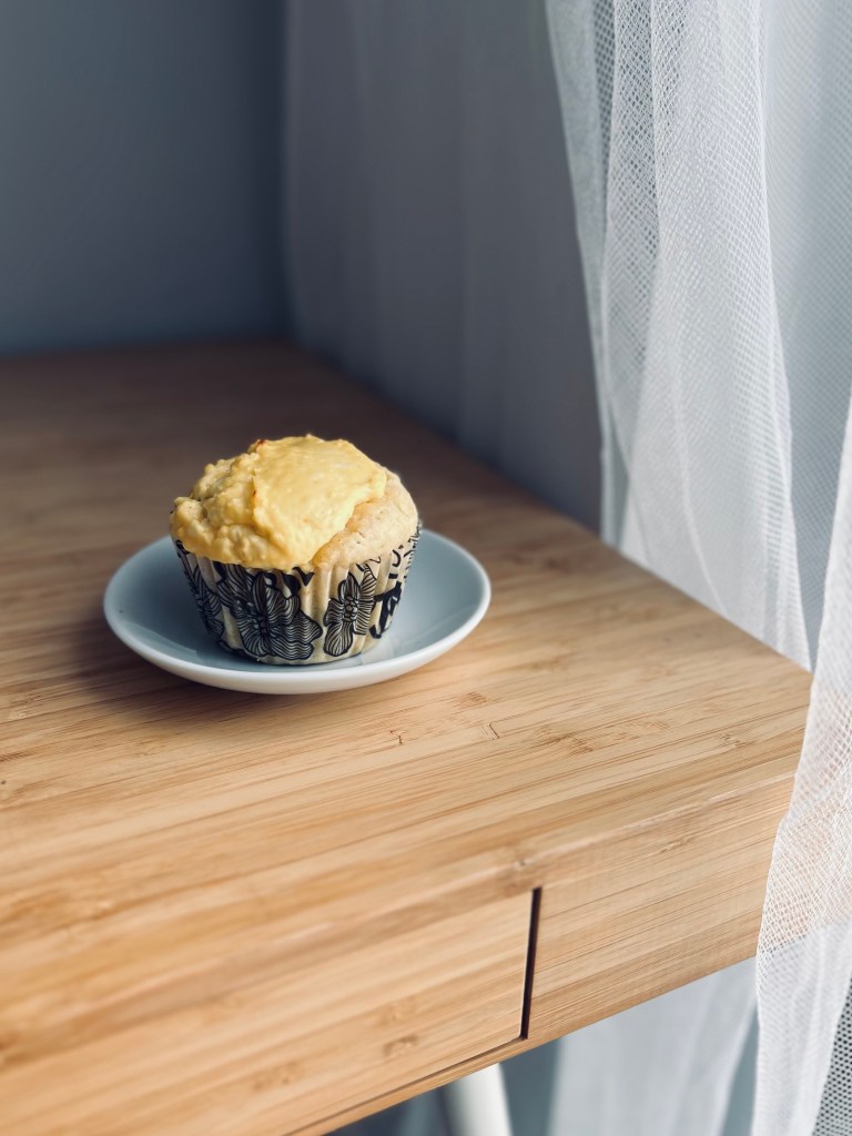 A freshly baked cupcake topped with creamy farmer's cheese frosting, placed on a small white plate on a wooden table next to a sheer curtain.